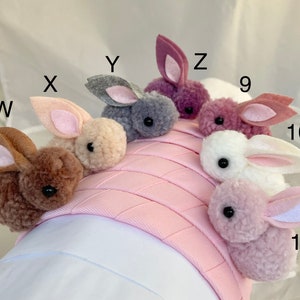 May include: A pink headband with nine fluffy bunny charms in various colors, including brown, gray, pink, and white. The charms are attached to the headband with a small loop.