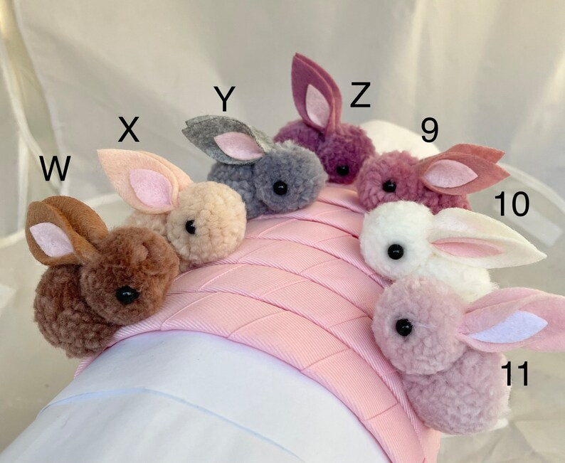 May include: A pink headband with nine fluffy bunny charms in various colors, including brown, gray, pink, and white. The charms are attached to the headband with a small loop.