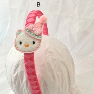 May include: Pink headband with a braided design and a white felt Hello Kitty character with a pink hat and bow. The headband is on a white surface.