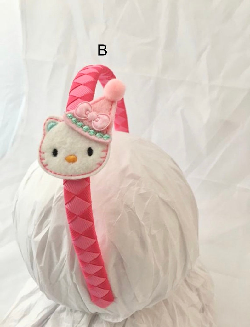 May include: Pink headband with a braided design and a white felt Hello Kitty character with a pink hat and bow. The headband is on a white surface.