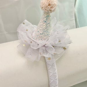 May include: White headband with a white tulle bow and a white glitter party hat with a white pom-pom on top. The tulle bow has gold stars on it.