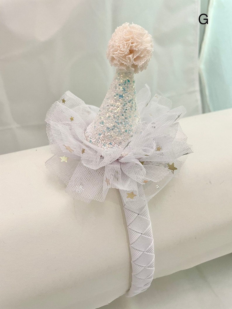 May include: White headband with a white tulle bow and a white glitter party hat with a white pom-pom on top. The tulle bow has gold stars on it.