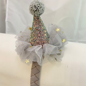 May include: A gray headband with a pink and silver glitter party hat and a white tulle bow with gold stars. The headband is made of a woven material.