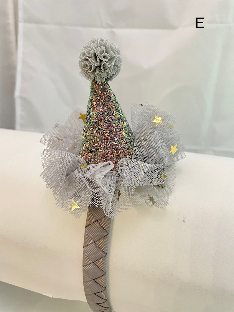 May include: A gray headband with a pink and silver glitter party hat and a white tulle bow with gold stars. The headband is made of a woven material.