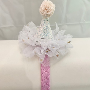 May include: A pink headband with a white tulle bow and a white glitter cone hat with a white pom-pom on top. The hat has gold glitter stars on it.
