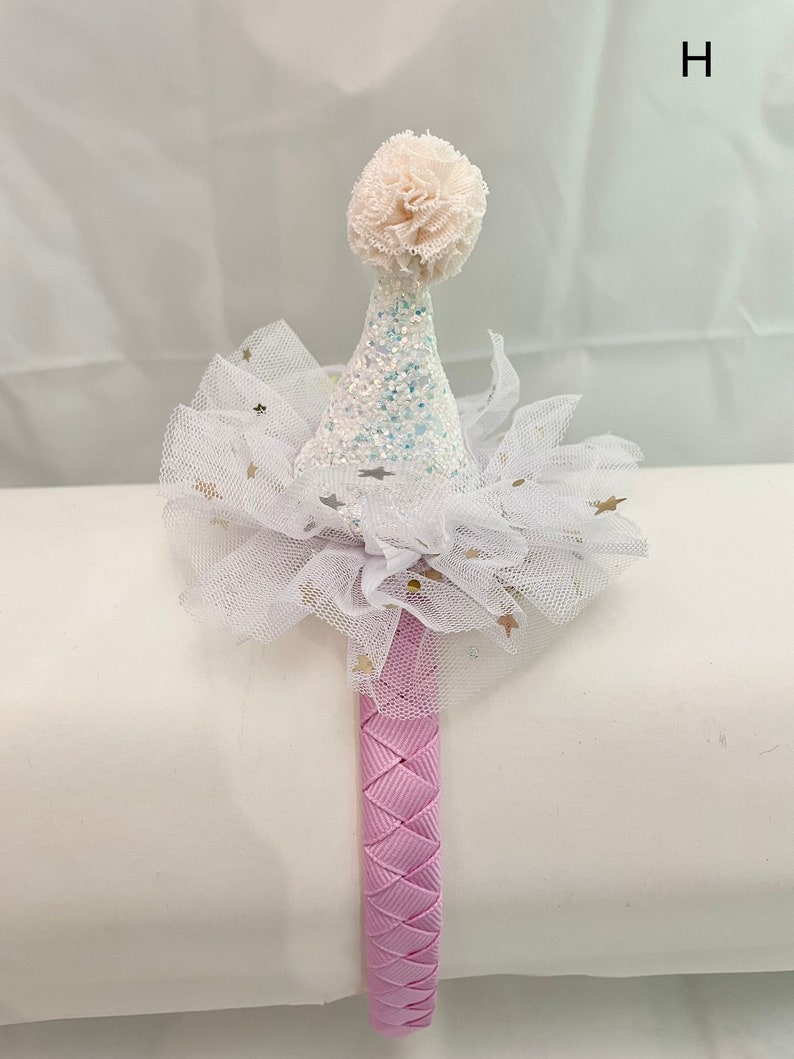May include: A pink headband with a white tulle bow and a white glitter cone hat with a white pom-pom on top. The hat has gold glitter stars on it.