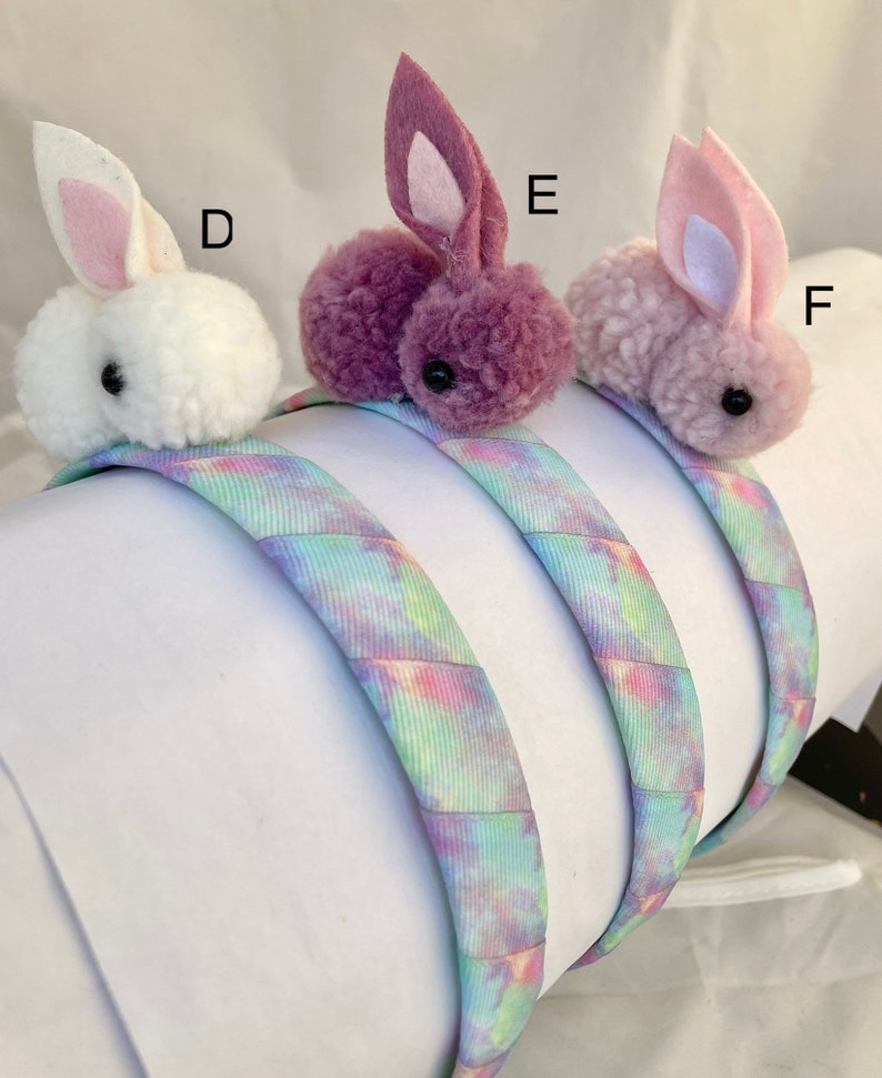 May include: Three headband hair accessories with fuzzy bunny charms. The headbands are tie-dye patterned in shades of blue, pink, and purple. The bunny charms are white, purple, and light pink.