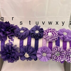 May include: Set of 9 purple and lavender fabric headbands with various flower and chevron designs. The headbands are labeled with the letters q, r, s, t, u, v, w, x, y, and z.