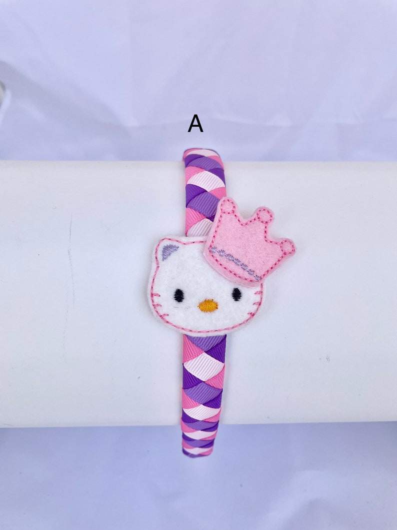 May include: A purple and pink braided headband with a white felt Hello Kitty character wearing a pink crown.