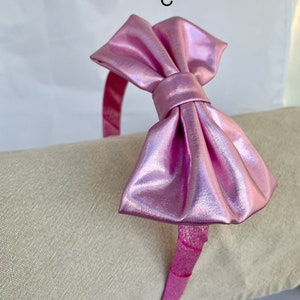 May include: A pink metallic headband with a large bow. The headband is on a light beige surface.