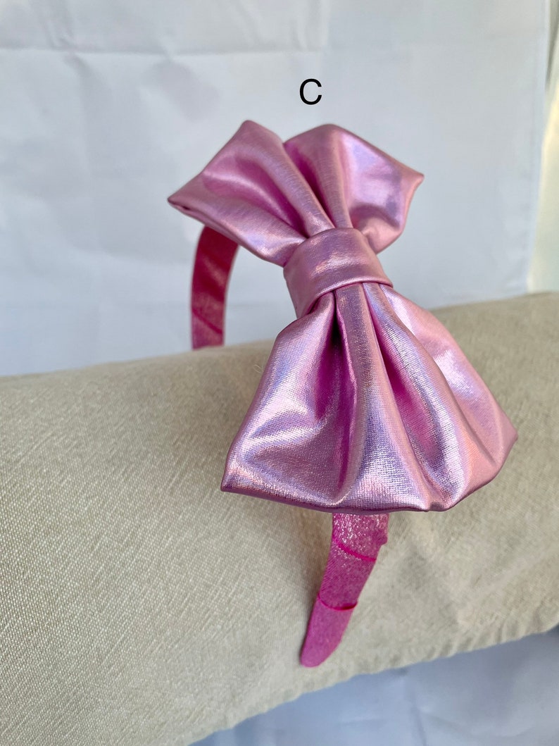 May include: A pink metallic headband with a large bow. The headband is on a light beige surface.