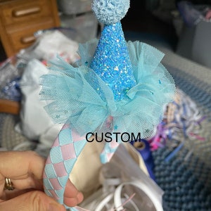 May include: A light blue headband with a pink and blue checkered ribbon. The headband has a light blue tulle bow and a light blue glitter party hat with a white pom-pom on top. The text "CUSTOM" is visible on the headband.