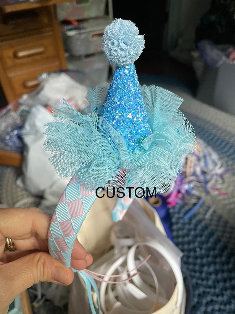 May include: A light blue headband with a pink and blue checkered ribbon. The headband has a light blue tulle bow and a light blue glitter party hat with a white pom-pom on top. The text "CUSTOM" is visible on the headband.