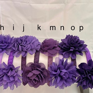 May include: Set of eight purple fabric flower headbands. The headbands are made of a soft, stretchy material and are decorated with a variety of purple fabric flowers.