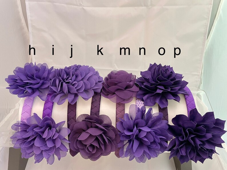 May include: Set of eight purple fabric flower headbands. The headbands are made of a soft, stretchy material and are decorated with a variety of purple fabric flowers.