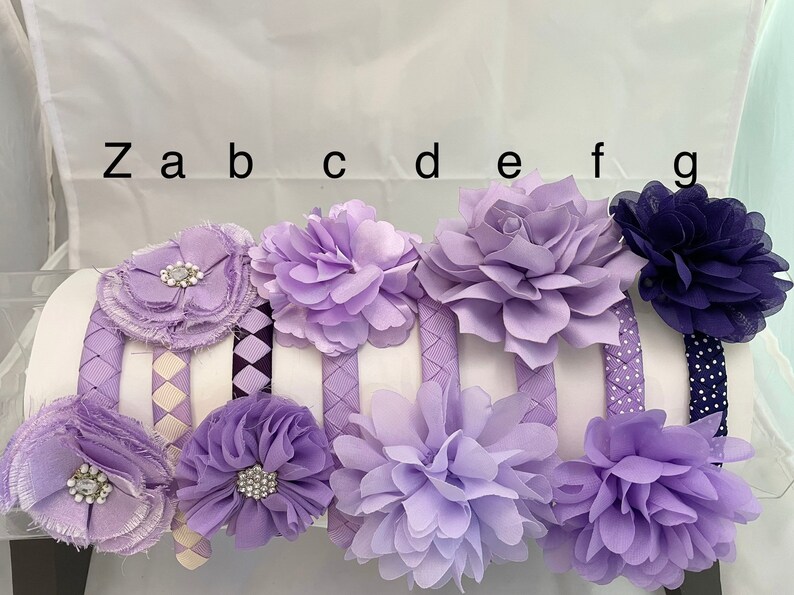 May include: Set of eight purple and navy blue headbands with various flower embellishments. The headbands have different patterns, including polka dots, plaid, and woven.