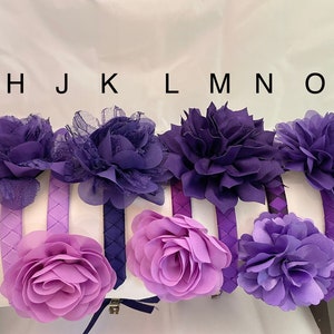 May include: Six purple and lavender fabric flower headbands with braided fabric bands. The headbands are arranged in a row with the letters H, J, K, L, M, N, and O above them.