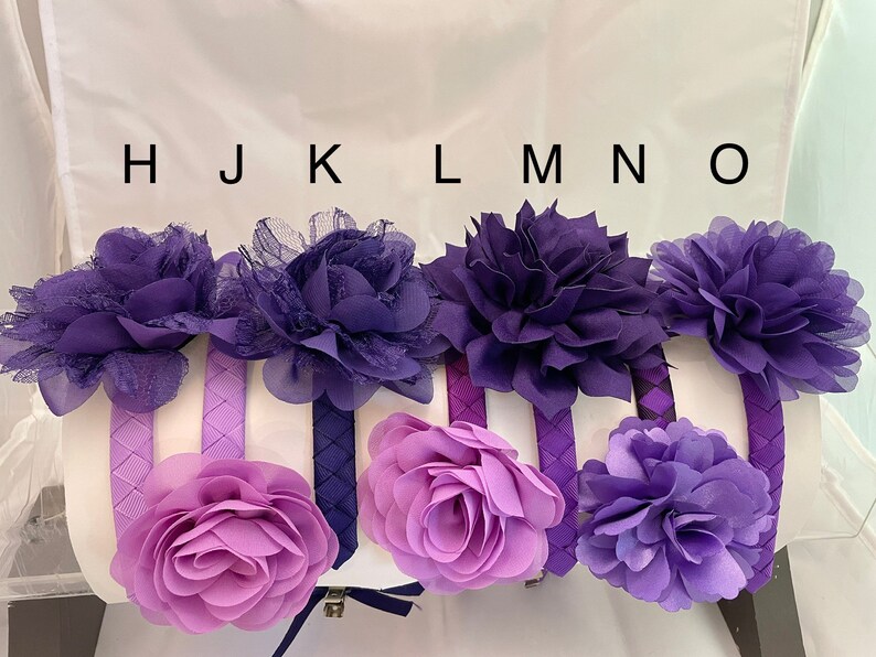 May include: Six purple and lavender fabric flower headbands with braided fabric bands. The headbands are arranged in a row with the letters H, J, K, L, M, N, and O above them.