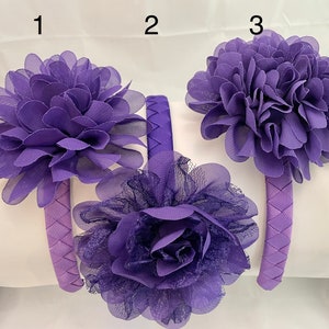May include: Three purple fabric headbands with different flower designs. The headbands are made of a woven fabric and have a purple flower on top. The flowers are made of fabric and have a layered look.