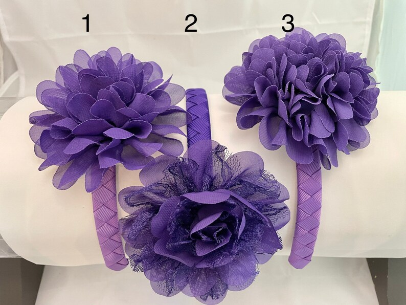 May include: Three purple fabric headbands with different flower designs. The headbands are made of a woven fabric and have a purple flower on top. The flowers are made of fabric and have a layered look.