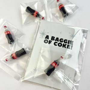 Puede incluir: Miniaturas de botellas de Coca-Cola en bolsas de plástico transparente. Cada botella es negra con una etiqueta roja y blanca y una tapa roja. La imagen incluye una tarjeta con el texto "A baggie of coke!" y gráficos de estrellas.