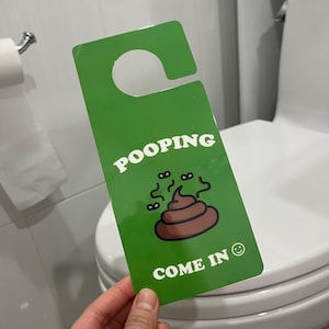 May include: Green door hanger with the word "POOPING" in white, a cartoon poop graphic, and the words "COME IN" with a smiley face. The hanger is held in front of a toilet.