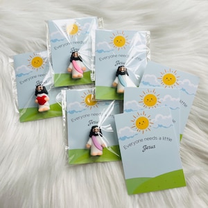 May include: Miniature Jesus figurines with cards. Each card features a smiling sun, clouds, and the text "Everyone needs a little Jesus." The figurines are in various poses, with robes in white, pink, and blue.