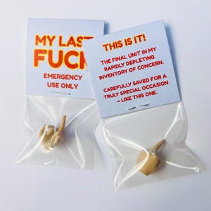 Op de afbeelding: Twee kleine plastic zakjes met elk een miniatuurhand die de middelvinger opsteekt. Elk zakje heeft een witte kaart met rode en oranje tekst. E&eacute;n kaart leest "MY LAST FUCK EMERGENCY USE ONLY." De andere leest "THIS IS IT!" en andere tekst.