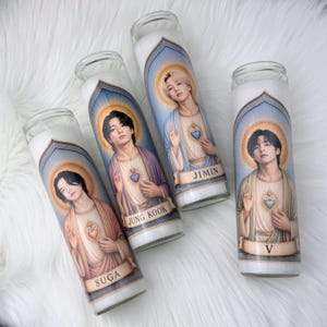 May include: Four cylindrical glass prayer candles featuring printed illustrations of individuals in robes with heart emblems. Each candle label displays a name: Suga, Jung Kook, Jimin, and V, set within an arched frame against a blue and gold background.