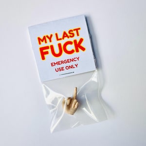 以下が含まれることがあります： ユーモラスなアイテム。中指を立てた小さな手は、透明なビニール袋に入っています。上のカードには、赤い文字に黄色の縁取りで「MY LAST FUCK」と書かれており、その下に「EMERGENCY USE ONLY」と書かれています。