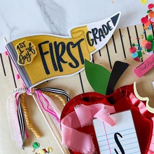 May include: A collection of back-to-school decorations, including a red apple-shaped plate, a banner that says "First Grade", and confetti. The apple plate has a small notepad with the letter "S" on it. The banner is yellow and white.
