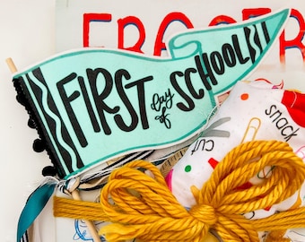 以下が含まれることがあります： 「First Day of School」の文字が入ったターコイズ色のペナント、黄色の糸の弓、そして「Libby」という名前の弓型のネームタグが特徴の、新学期をテーマにしたギフトセットです。学校関連の様々なアイテムが含まれています。