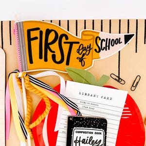 May include: A colorful back-to-school themed scrapbook page. The page features a yellow pennant with the words "First Day of School", a red apple, a library card, and a composition book with the name "Hailey". Ribbons and paperclips add to the design.