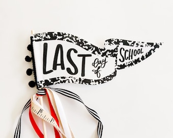 以下が含まれることがあります： 「LAST day of SCHOOL」の文字が黒で書かれた装飾的な旗。旗は白と黒の模様の縁取り、黒いポンポン、カラフルなリボンが付いています。木製のダボが旗を支えています。