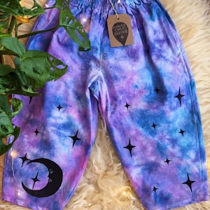 May include: Tie-dye trousers in blue, purple, and pink, featuring black star and crescent moon designs. They have an elasticated waist and a tag reading "Flower Child Bazaar". The trousers are shown on a fluffy, cream-coloured surface.