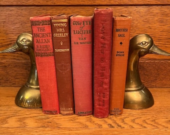 May include: A row of six vintage hardcover books with red, brown, and orange covers. The books are arranged on a wooden shelf with two brass duck bookends on either end.