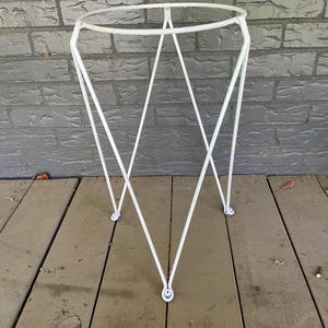 May include: A white metal plant stand with a circular top and a geometric design. The stand has three legs and is approximately 24 inches tall.