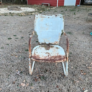 May include: A white metal chair with rust showing through the paint. The chair has a curved back and a simple design. It is sitting on a patch of brown grass.