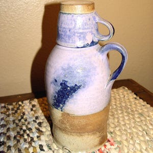 May include: A handmade ceramic pitcher and lid set. The pitcher is a light purple with blue and brown accents. The lid is a small cup with a handle. The pitcher has a handle. The set is on a woven mat.