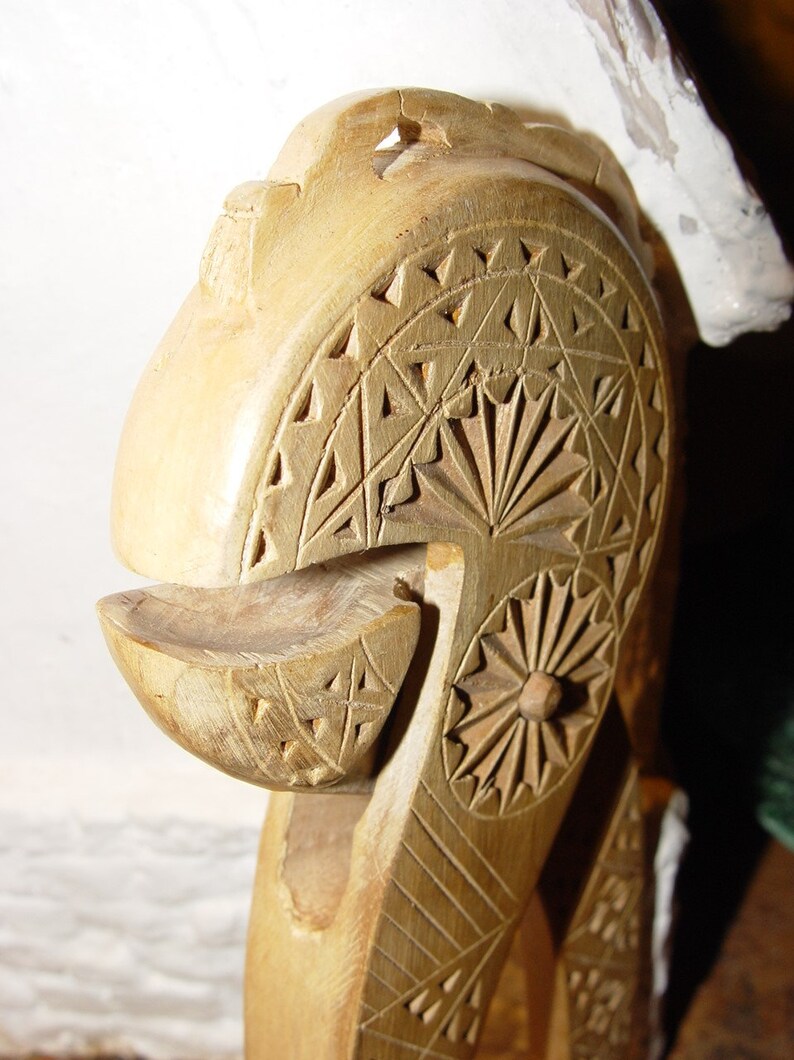 May include: A wooden nutcracker carved with intricate geometric patterns. The nutcracker is made of light brown wood and has a rounded head with a small opening for cracking nuts.