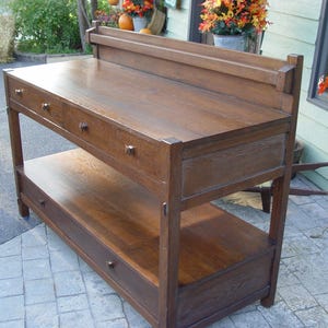 May include: A dark brown wooden console table with three drawers on the top level and a shelf with a drawer below. The table has a rectangular shape and is made of wood. The table is approximately 48 inches wide, 18 inches deep, and 36 inches tall.