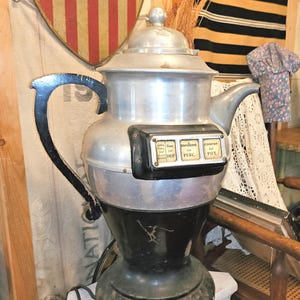 May include: A vintage aluminum coffee percolator with a black handle and base. The front features a black rectangular panel with four white labels reading extra fine for glass maker, fine for drip, medium for perc, and coarse for pot.