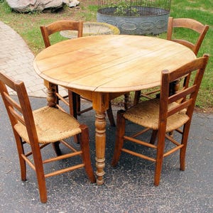 May include: A round wooden dining table with four matching chairs. The table has a light brown finish and a pedestal base. The chairs have woven seats and wooden frames. The set is outdoors on a paved surface.