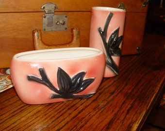May include: Two pink ceramic vases with black floral designs. The vases are on a wooden surface.