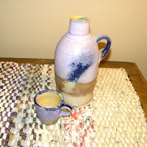 May include: A handmade ceramic jug and small mug. The jug is light purple with a blue design and a brown base. The mug is light purple with a blue handle. Both are on a woven rug with a mix of colors.
