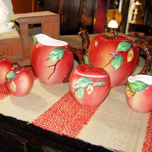 May include: A red apple-shaped tea set with green leaves and brown branch handles. The set includes a teapot, creamer, sugar bowl, and two salt and pepper shakers. The set is displayed on a red and white patterned tablecloth.