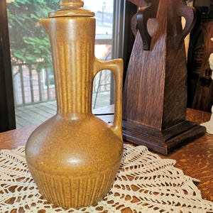 May include: A brown ceramic pitcher with a speckled finish, featuring a rounded base, a tall neck with vertical ribbing, a side handle, and a matching lid. The pitcher has a distinct spout and a uniform earthy tone throughout its entire structure.
