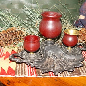 May include: A vintage cast iron tobacco pipe stand with three red wooden bowls. The stand has an ornate design and is sitting on a colorful rug.