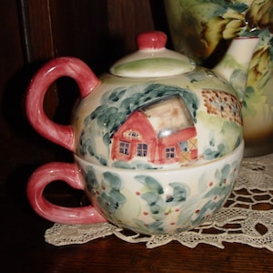 May include: A two-piece ceramic teapot and teacup set with a red, white, and green floral design. The teapot features a painted image of a red house with white trim and a green roof. The teacup has a green and white floral design.