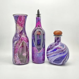 May include: Three decorative glass bottles with purple and white marbled designs. The bottles have glitter accents in pink and silver. One bottle has a spout, and another has a wooden stopper. The bottles are on a white background.